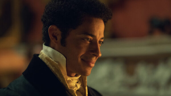"A man dressed in historical formal attire smiles warmly in a softly lit, elegant interior, likely from a period drama setting."
