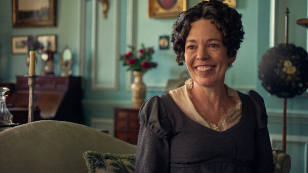 "A smiling woman in period costume sits on a couch in a vintage, elegant living room with teal walls, antique furniture, framed art, and a flower vase in the background."